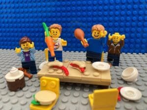 A family trying to eat dinner at the table but two children are dancing on top. Plates and food are all over the floor.