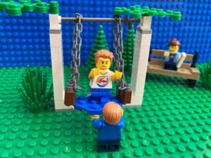 Child pushing his brother on a swing while he takes the risk to not hold on with his hands. Mum looking horrified in the background