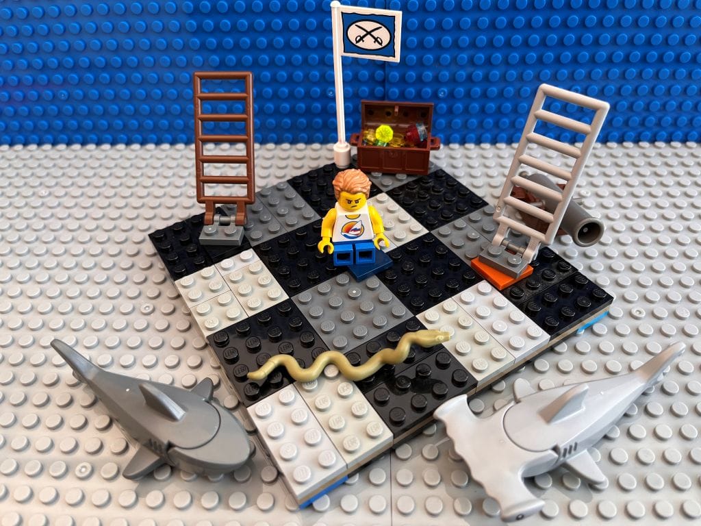 Cross child sitting in the middle of a large snakes and ladders board game with toy sharks, treasure chests and a pirate flag